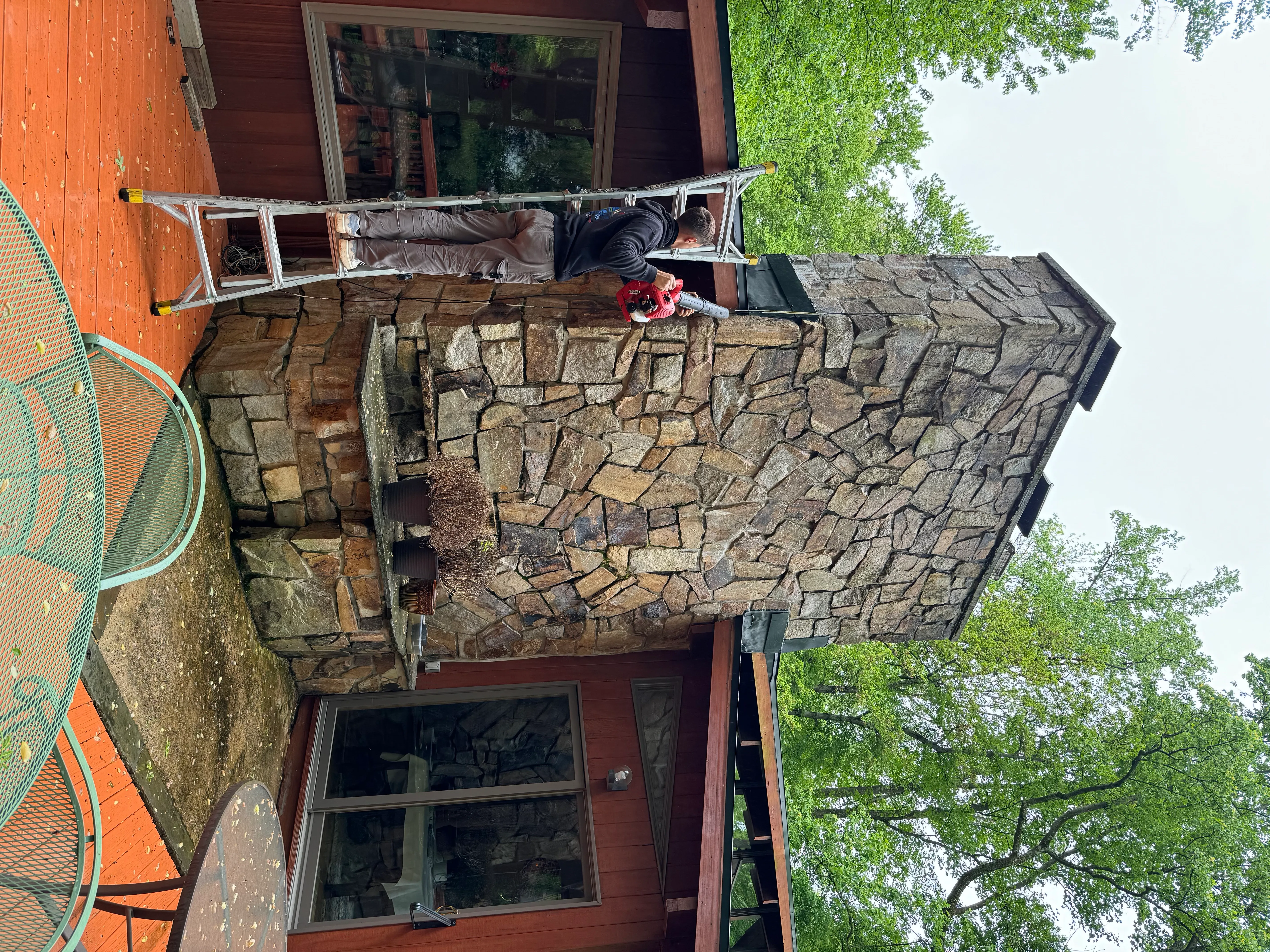 Roofer on a ladder repairing the tall stone chimney above the cedar home and back deck.