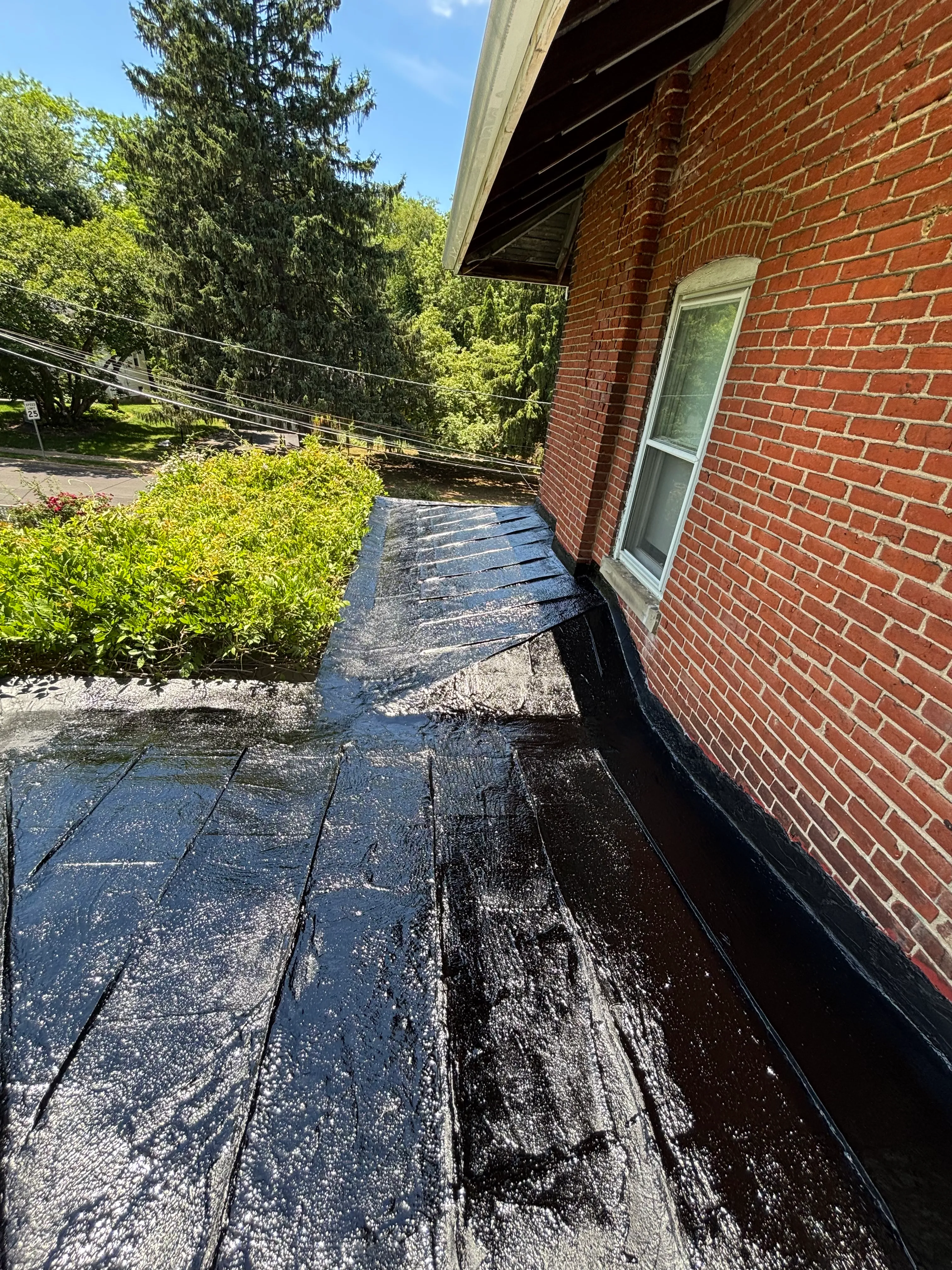 Parapet freshly coated in wet, glossy black roof coating, the brick house and a skylight behind it.