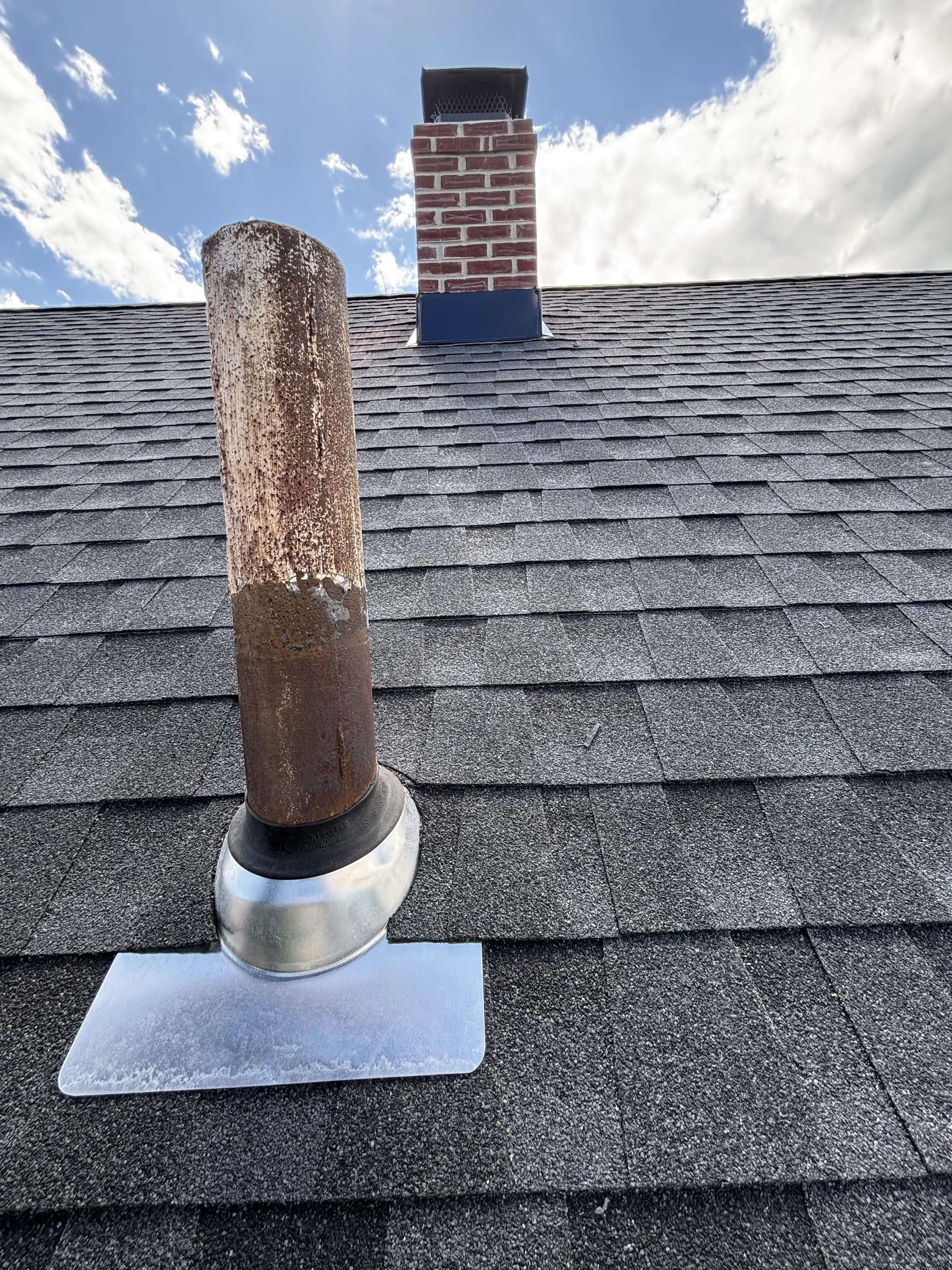 New vent-pipe flashing seated into the finished charcoal shingles, with the rebuilt brick chimney in the background.