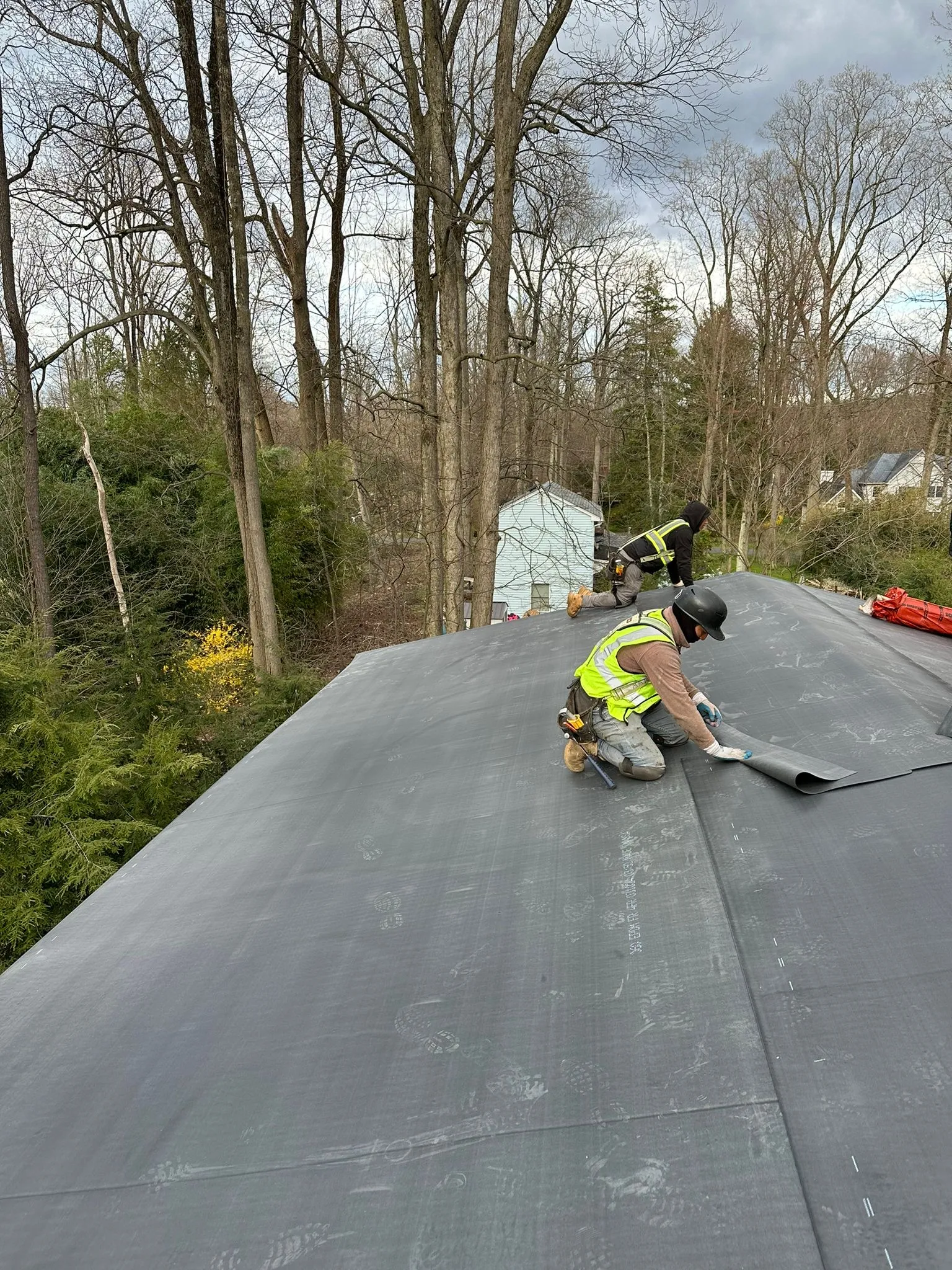 Roofer rolling out black EPDM membrane over the insulated deck, a second worker at the roof edge.